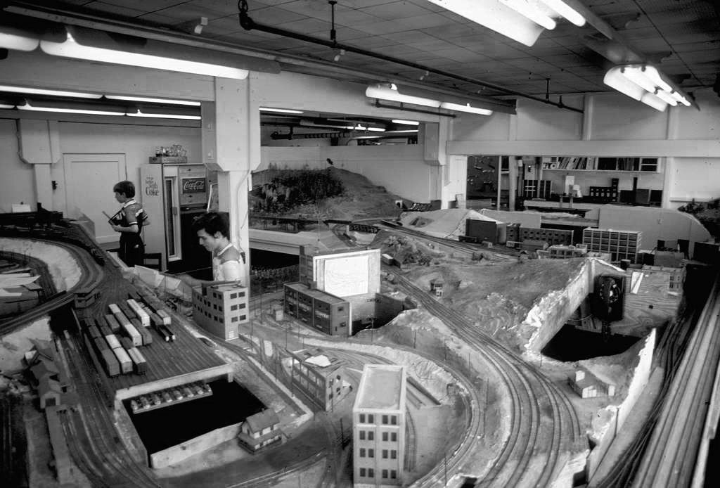 The meeting space of MIT's Tech Model Railroad Club in the 1960s, with a half finished model train track and students working on it.